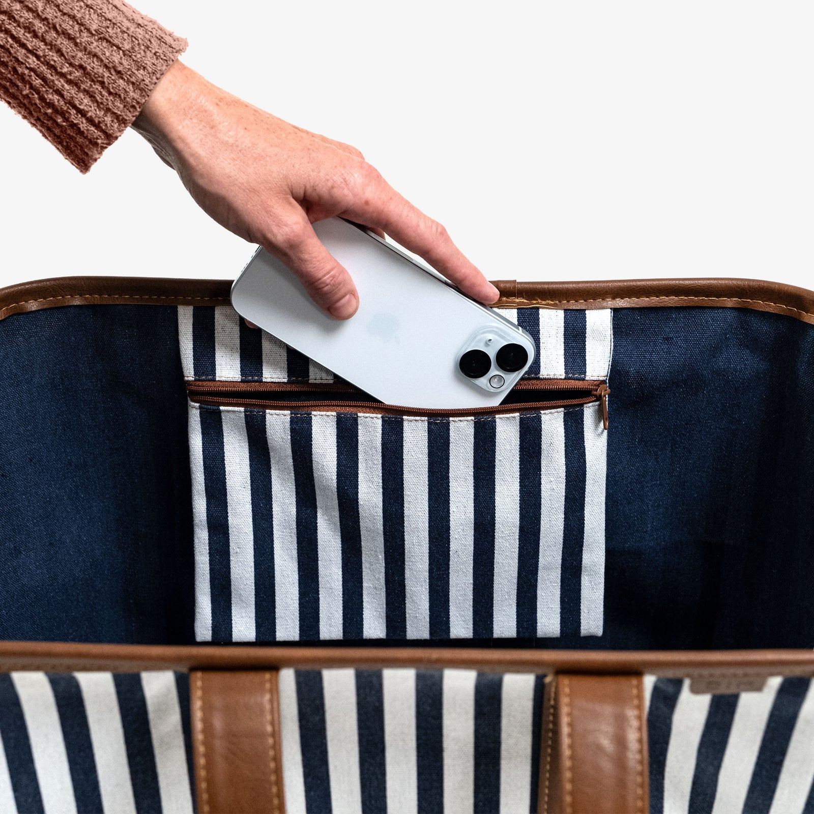 Hand placing a smartphone into a navy and white striped bag with brown leather trim.