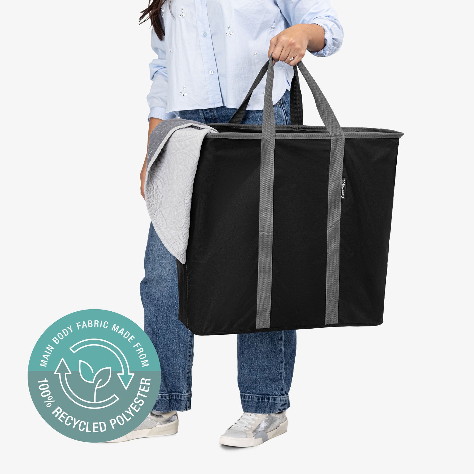 Person holding a black tote bag with gray straps on a white background, featuring a label indicating recycled polyester material.