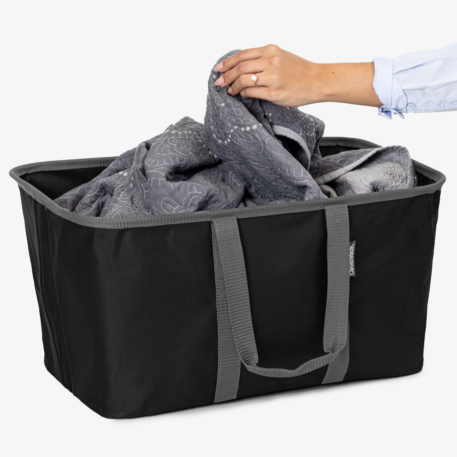 Person placing a folded blanket into a black laundry tote bag on a white background