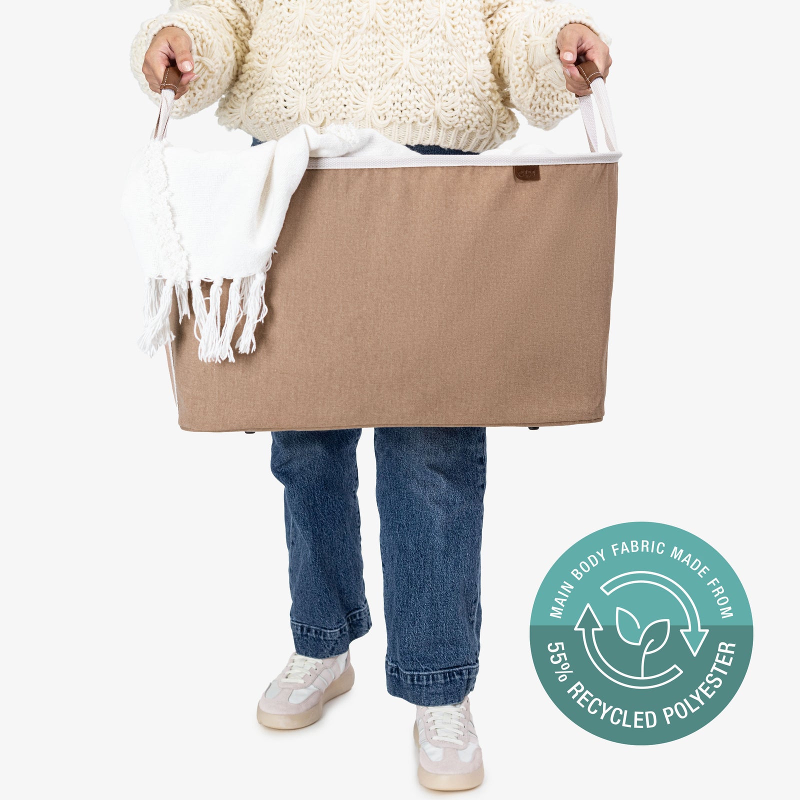 Person holding a beige collapsible laundry basket LUXE with a blanket, wearing jeans and white sneakers, on a white background.