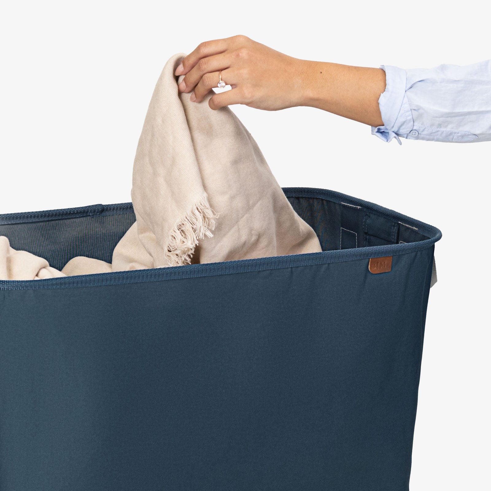 Person placing a beige towel into a navy blue laundry basket on a white background