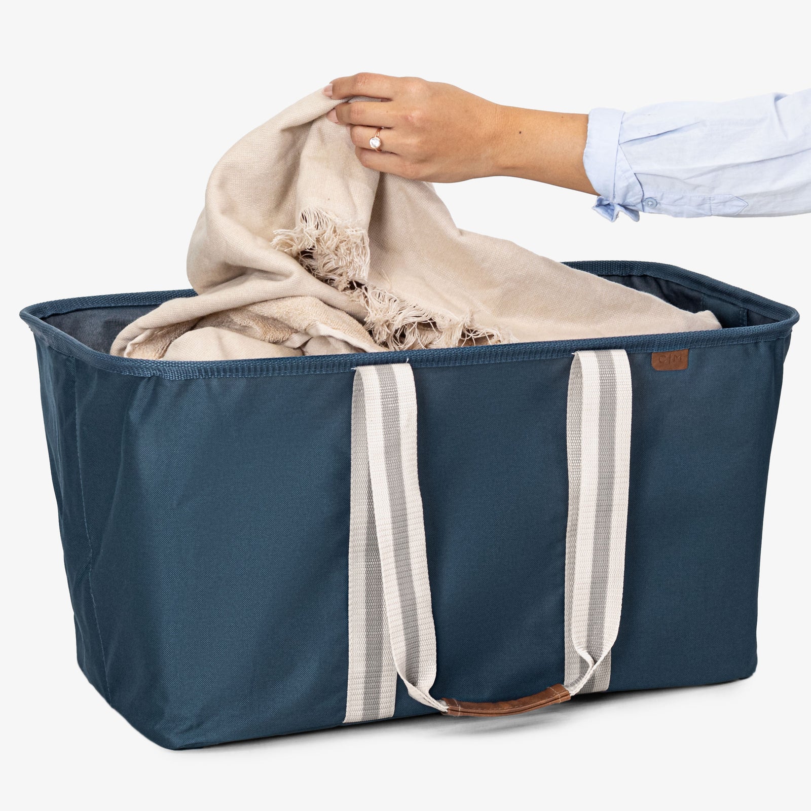 Blue laundry tote bag with white straps being filled with a beige blanket by a person wearing a light blue shirt.