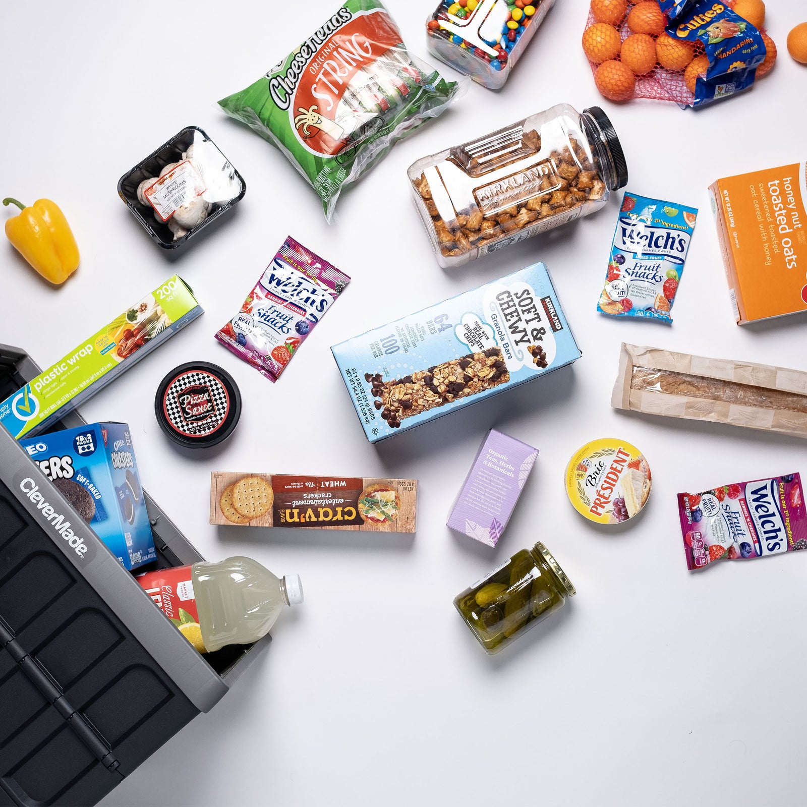 A gray and black collapsible storage bin with lid with assorted snacks and food items on a white surface