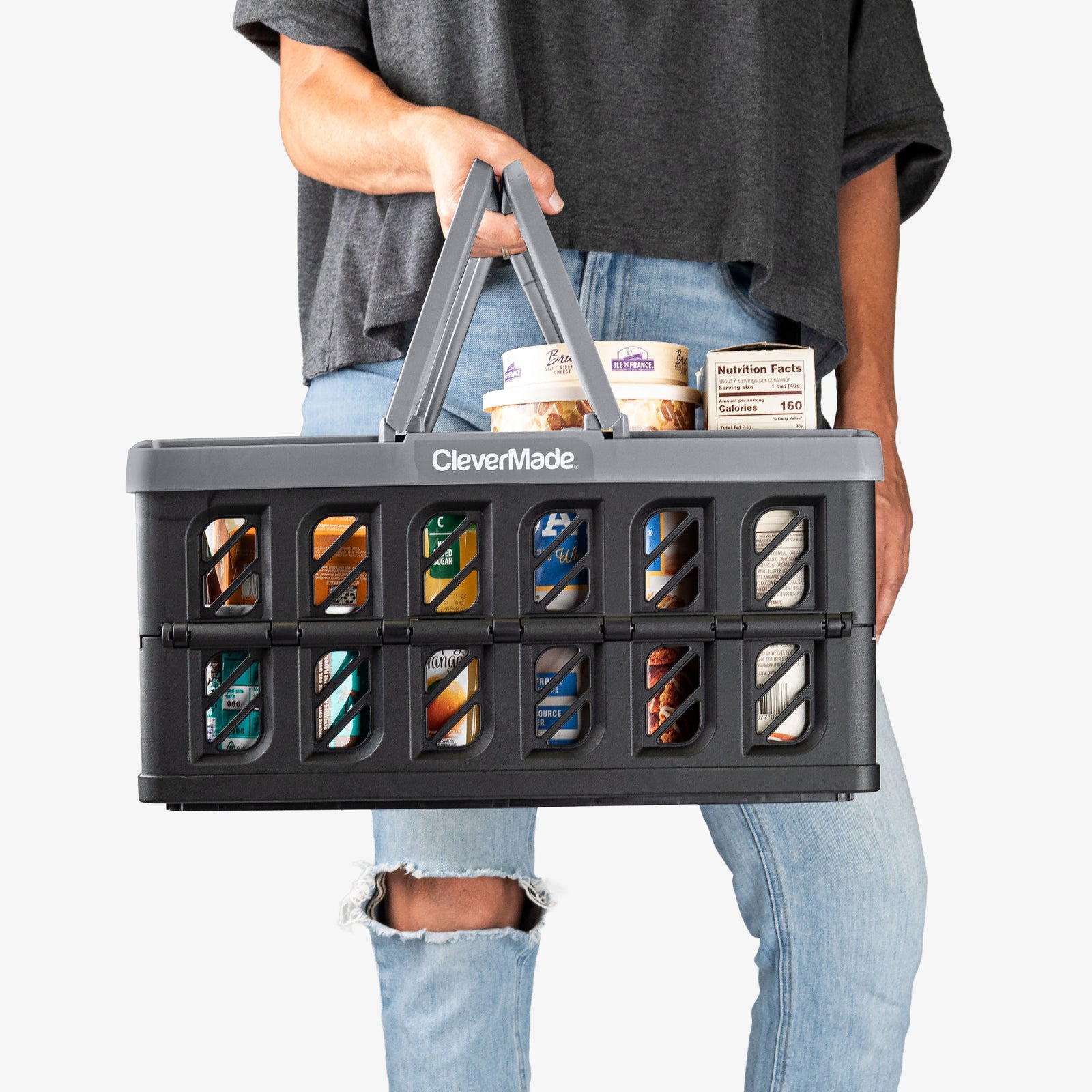 Person holding a CleverMade gray-black collapsible shopping basket with various items inside on a white background