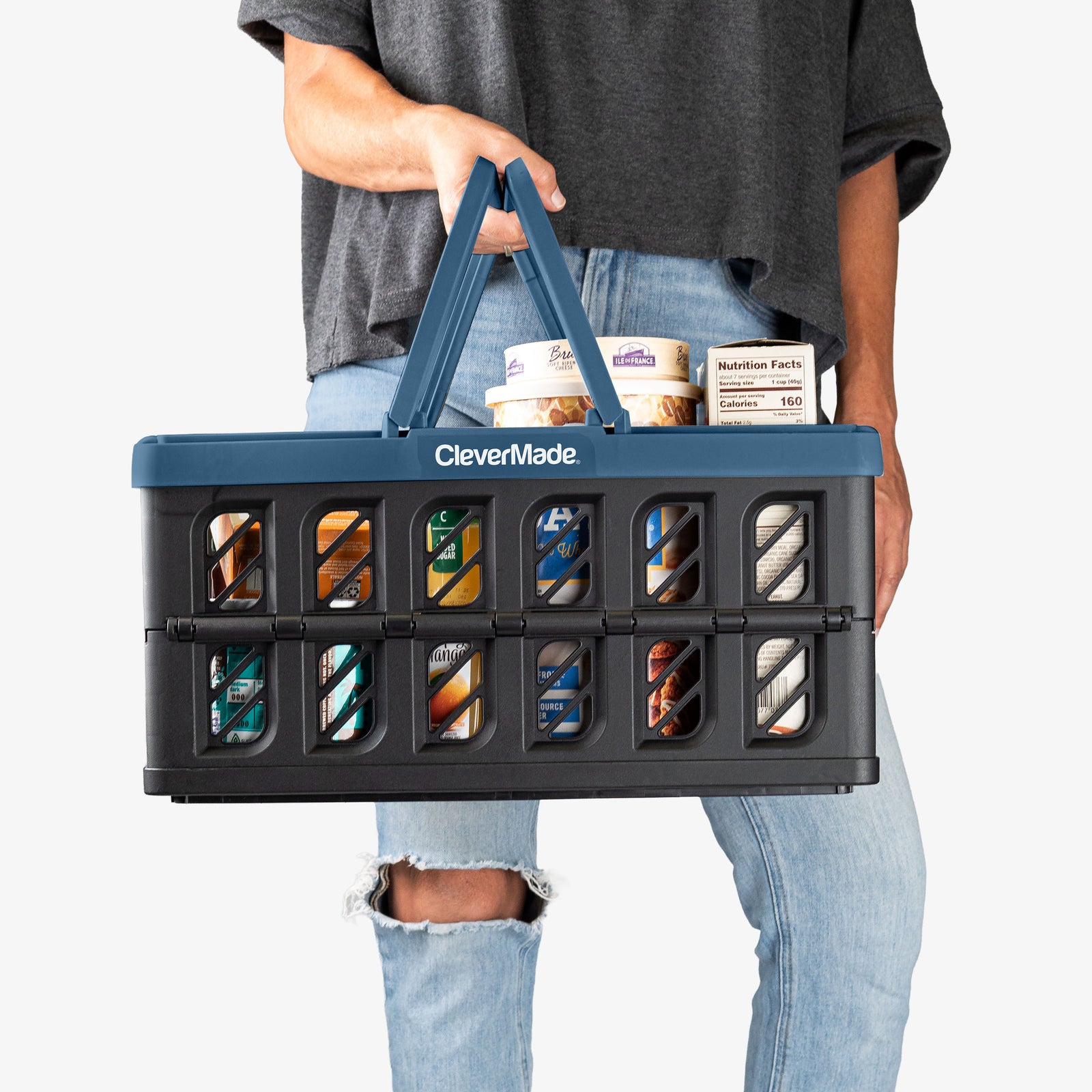 Person holding a CleverMade navy and black collapsible shopping basket with food items on a white background