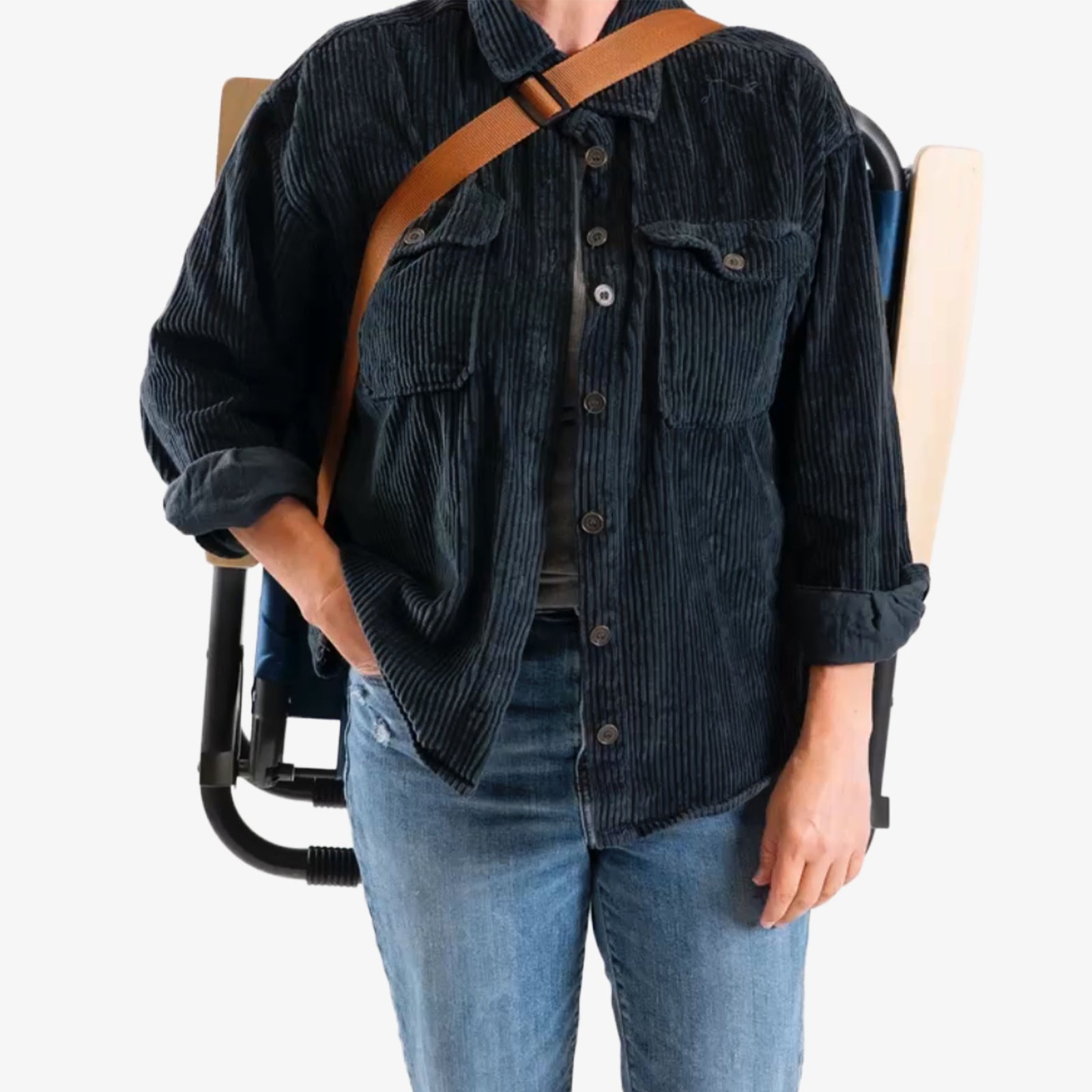 Person wearing a dark corduroy shirt and blue jeans carrying folded Kove outdoor chair with strap over shoulder