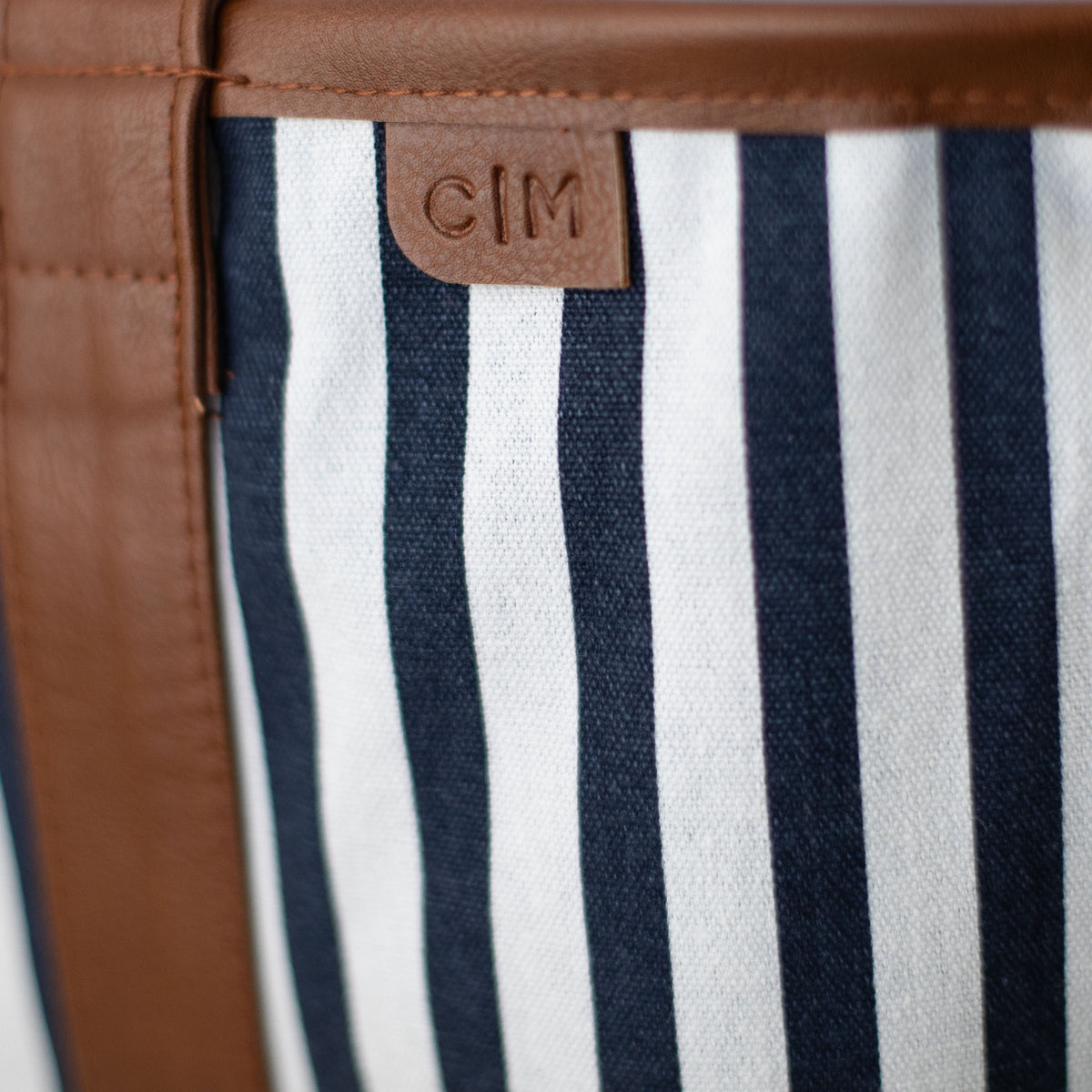 Close-up of a tote bag with striped fabric and brown leather trim, featuring the brand 'C|M'.
