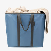 Blue laundry caddy with black handles filled with beige towels on a white background