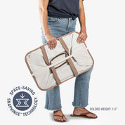 Person holding a folded beige and brown laundry tote LUXE with 'Space-Saving SnagHinge Technology' label.