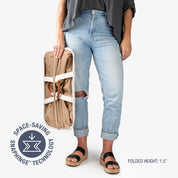 Person holding a folded beige laundry tote LUXE bag with space-saving snaphinge technology logo, wearing light blue jeans and black sandals on a white background.