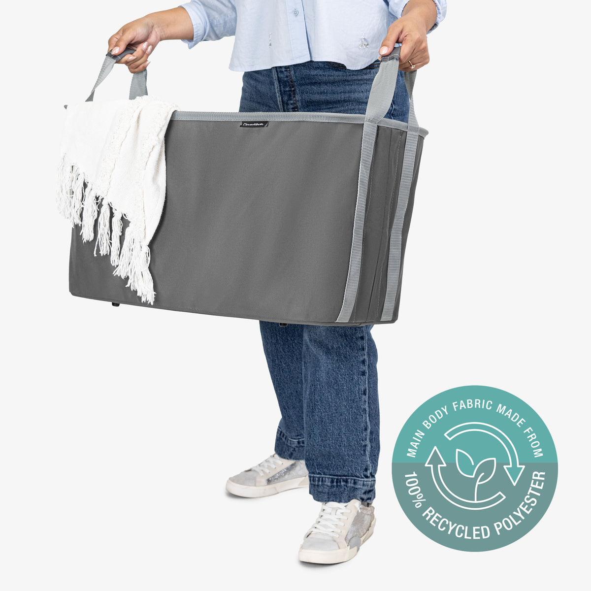 Person holding a large gray collapsible laundry basket with a white towel with tassel, on a white background with a circular label indicating 100% recycled polyester.