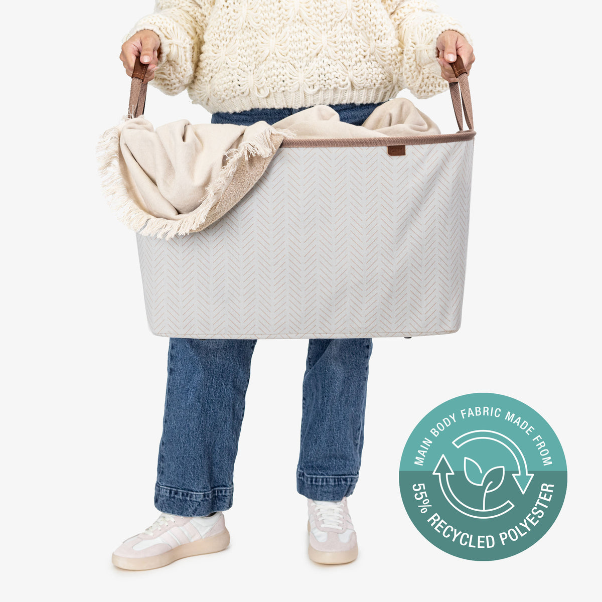 Person holding a large cream collapsible laundry basket LUXE
 with a blanket, wearing a white sweater and blue jeans, on a white background.
