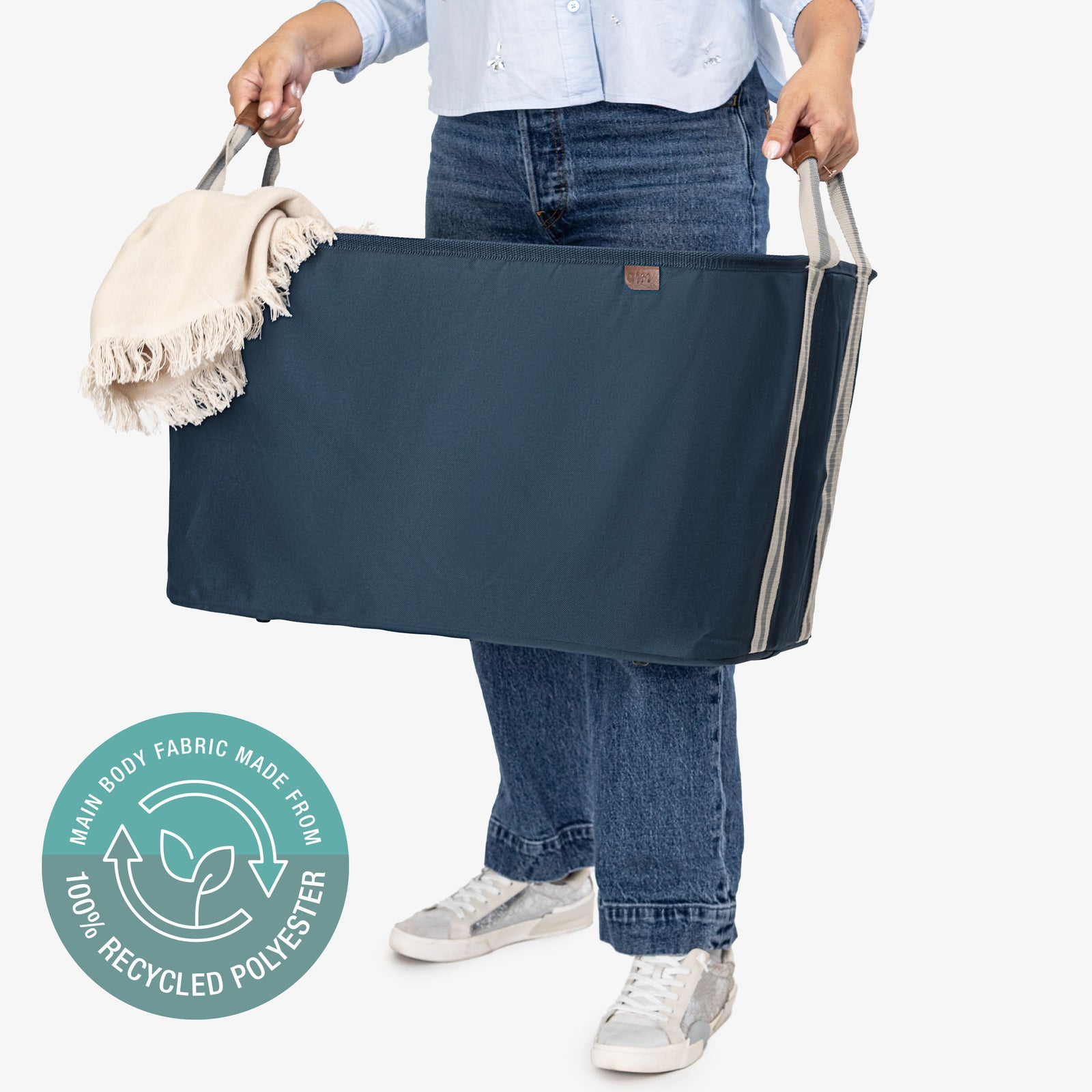Person holding a large navy collapsible laundry basket with a white towel with tassel, on a white background with a circular label indicating 100% recycled polyester.
