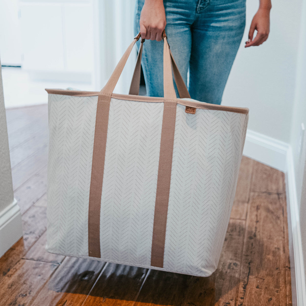 Person holding a large beige tote bag with brown straps on a wooden floor.