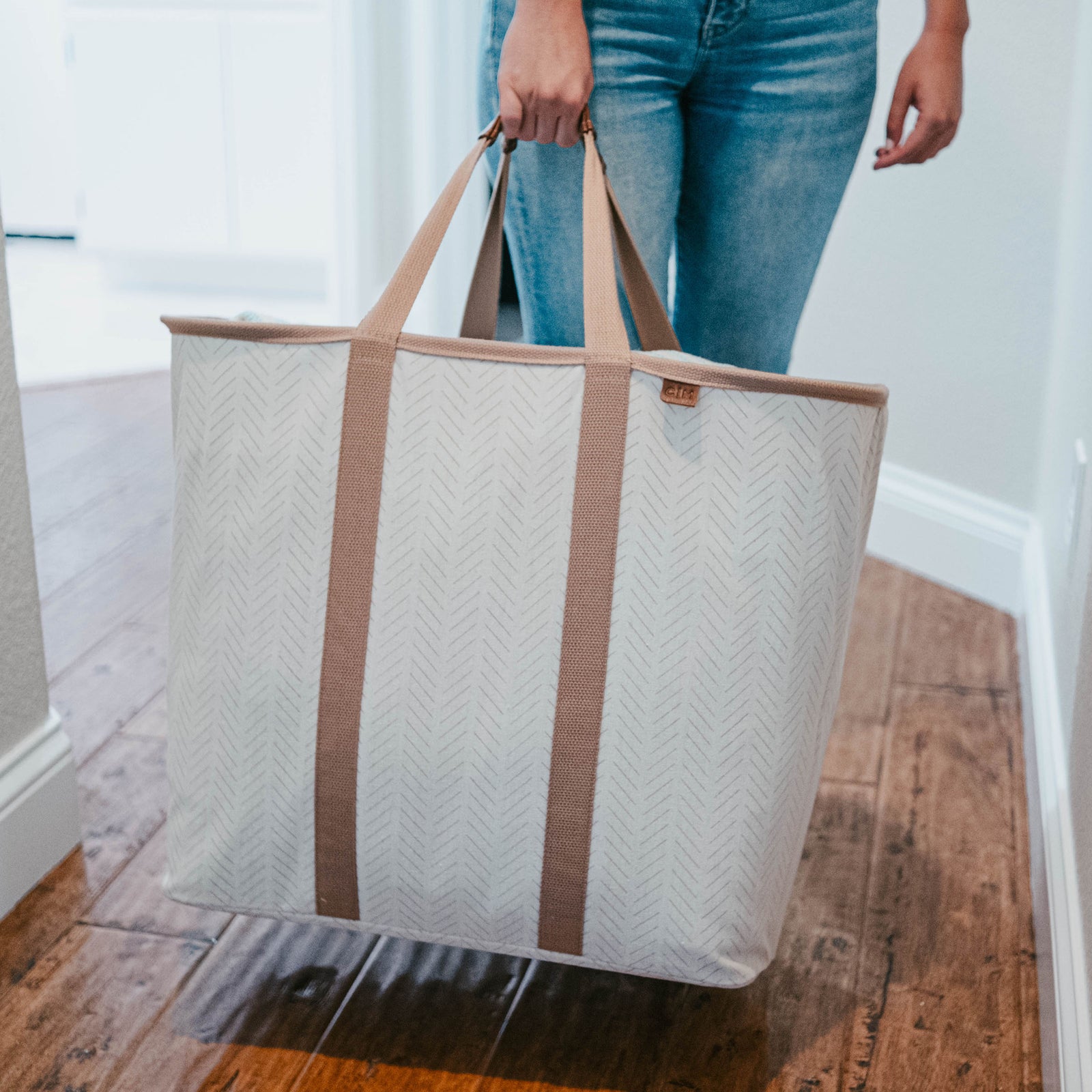 Person holding a large beige tote bag with brown straps on a wooden floor.