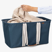 Blue laundry tote bag with white straps being filled with a beige blanket by a person wearing a light blue shirt.