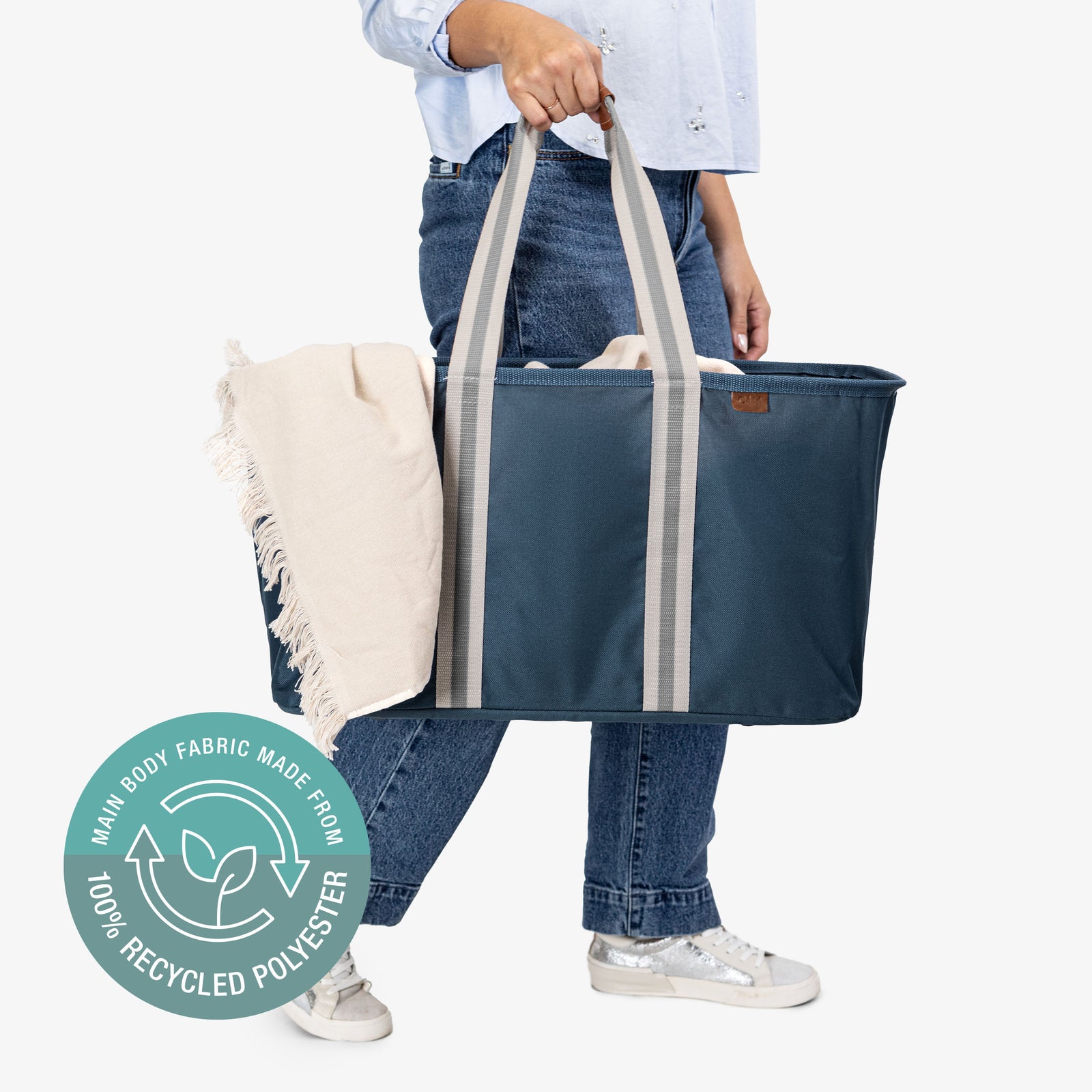 Person holding a blue tote bag with a blanket, wearing jeans and white sneakers, on a light gray background.