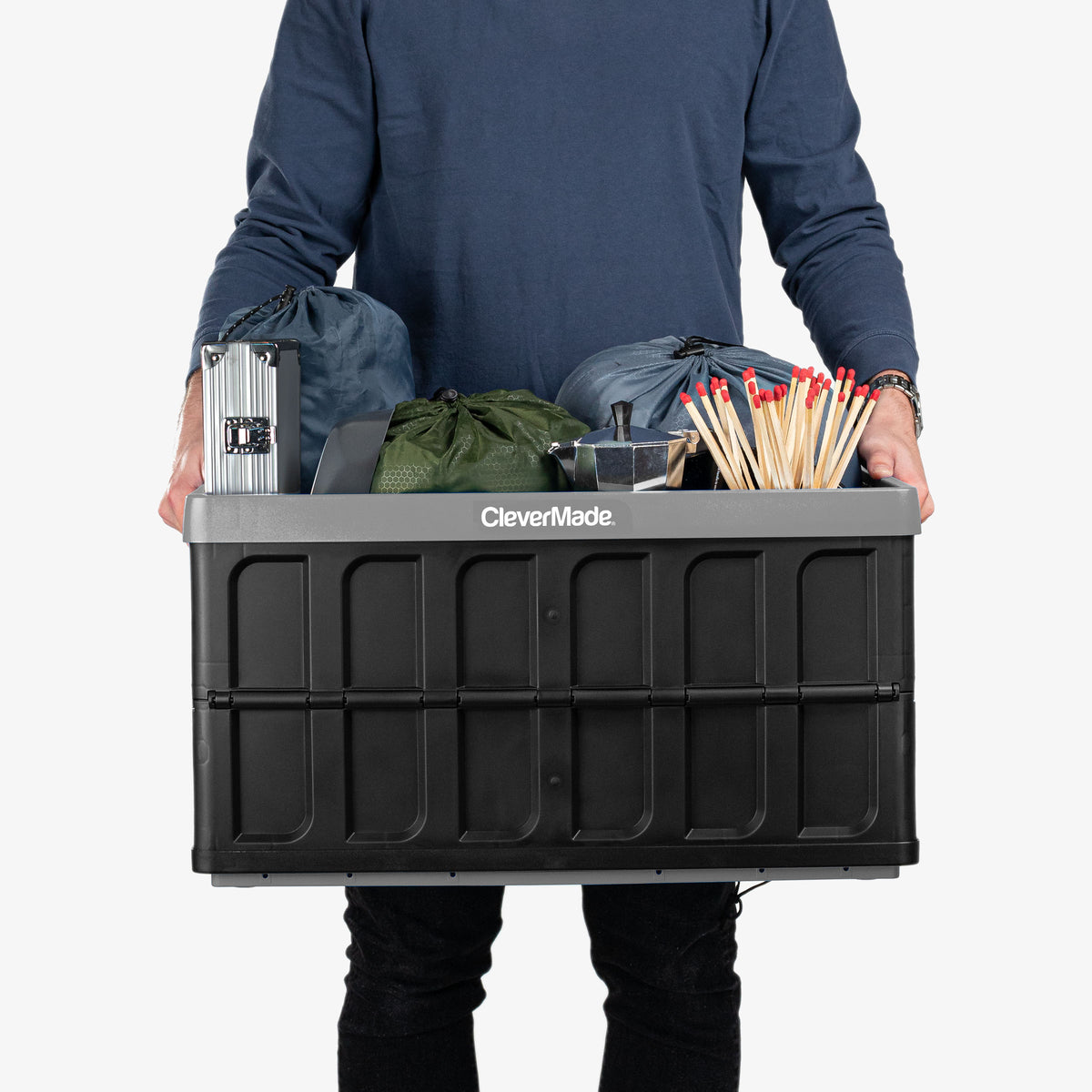 Person holding a CleverMade gray and black collapsible storage bin filled with various items on a white background