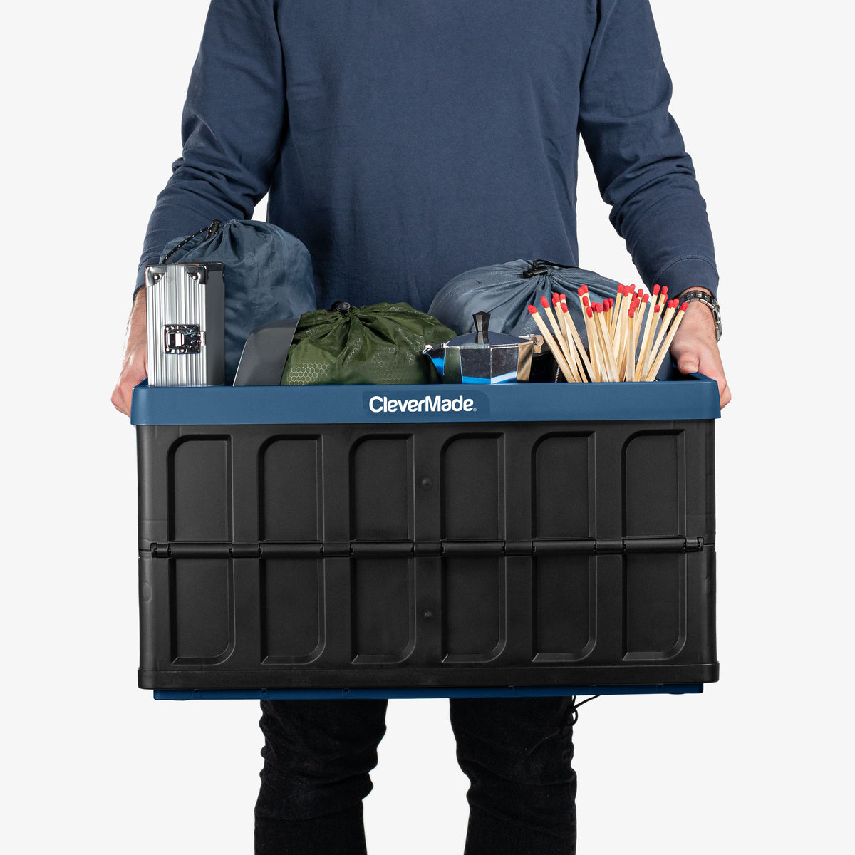 Person holding a CleverMade blue and black collapsible storage bin with lid filled with various items on a white background