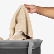Person placing a beige towel into a gray collapsible laundry tote on a white background