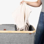 Person placing a beige blanket into a gray lidded hamper with a wooden handle.