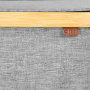 Close-up of a gray lidded hamper with a wooden edge and a brown leather patch with 'C|M' branding.