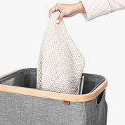 Person placing a white knitted blanket into a gray laundry basket with a wooden handle.