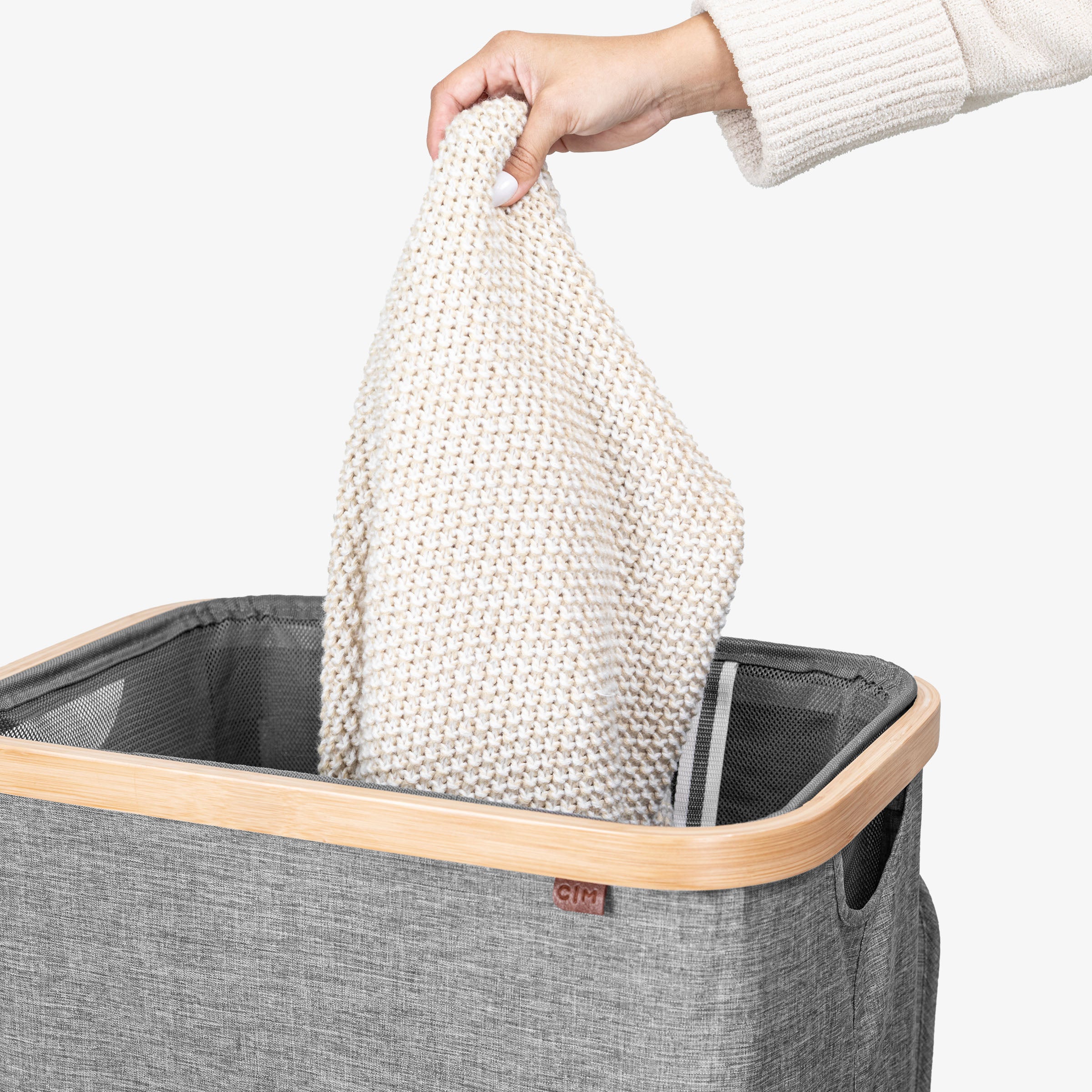 Person placing a white knitted blanket into a gray laundry basket with a wooden handle.
