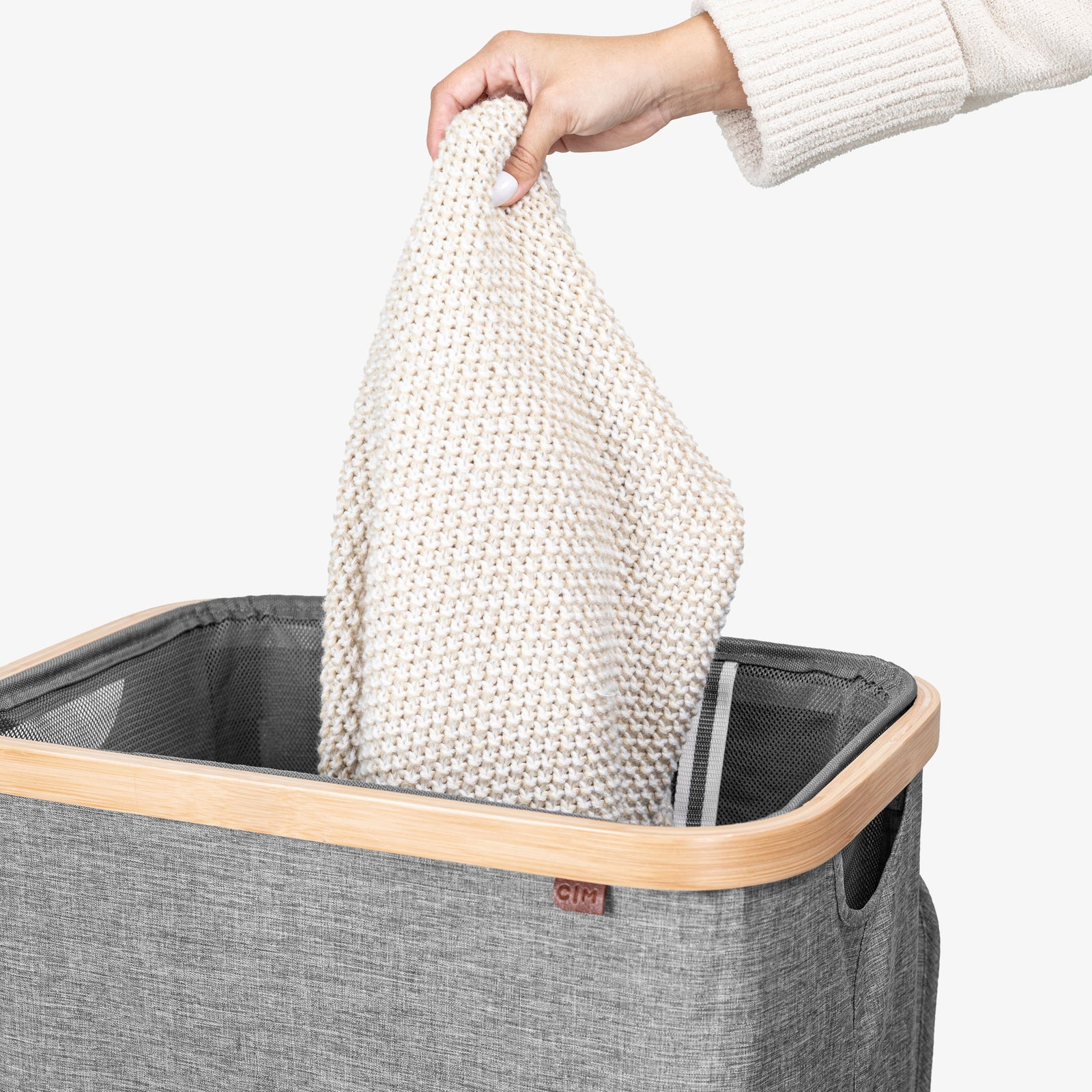 Person placing a white knitted blanket into a gray laundry basket with a wooden handle.