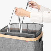 Person lifting a gray laundry basket with a mesh compartment and wooden rim.