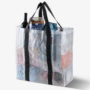 Clear reusable storage tote bag with black handles filled with groceries on a white background