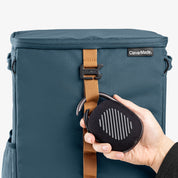 Blue backpack cooler bag with a brown strap and a black speaker held by a hand on a white background