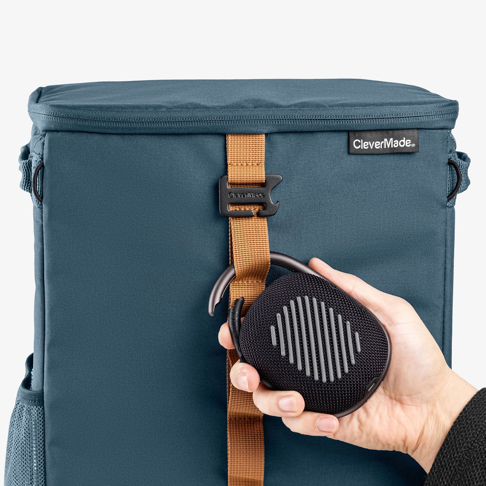 Blue backpack cooler bag with a brown strap and a black speaker held by a hand on a white background